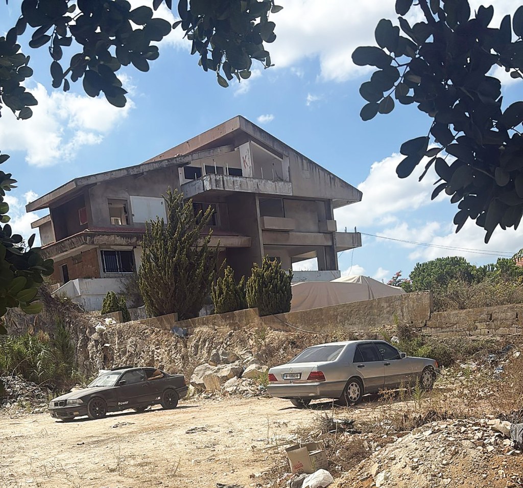 Ett stort övergivet hus står på en vildvuxen tomt med två bilar framför.